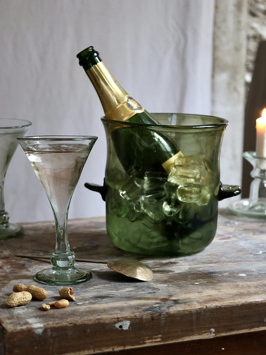 Bottle of champagne in a green glass ice bucket with a cocktail glass and peanuts on a wooden surface.