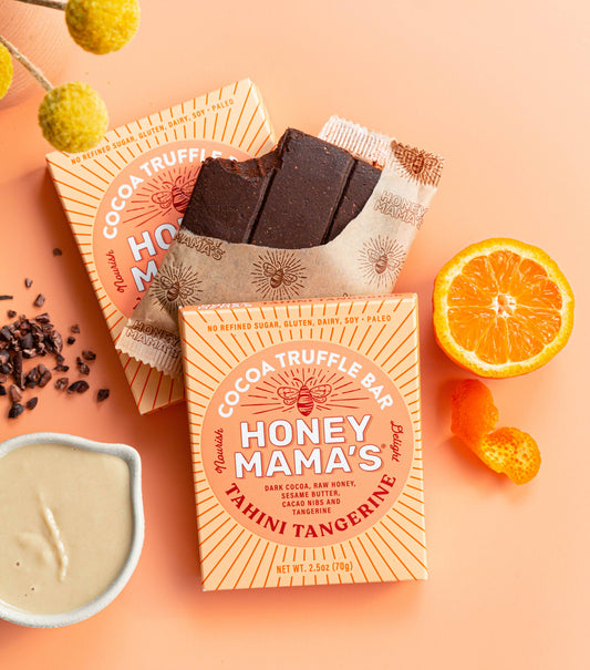 Honey Mama's Sesame Citrus Cocoa Truffle Bar. The packaging is orange and white with product information stating it is made from real foods only, paleo, refined sugar-free, dairy-free, and gluten-free. It's set up with a tangerine, sesame butter and cacao nibs next to the packaged bars.