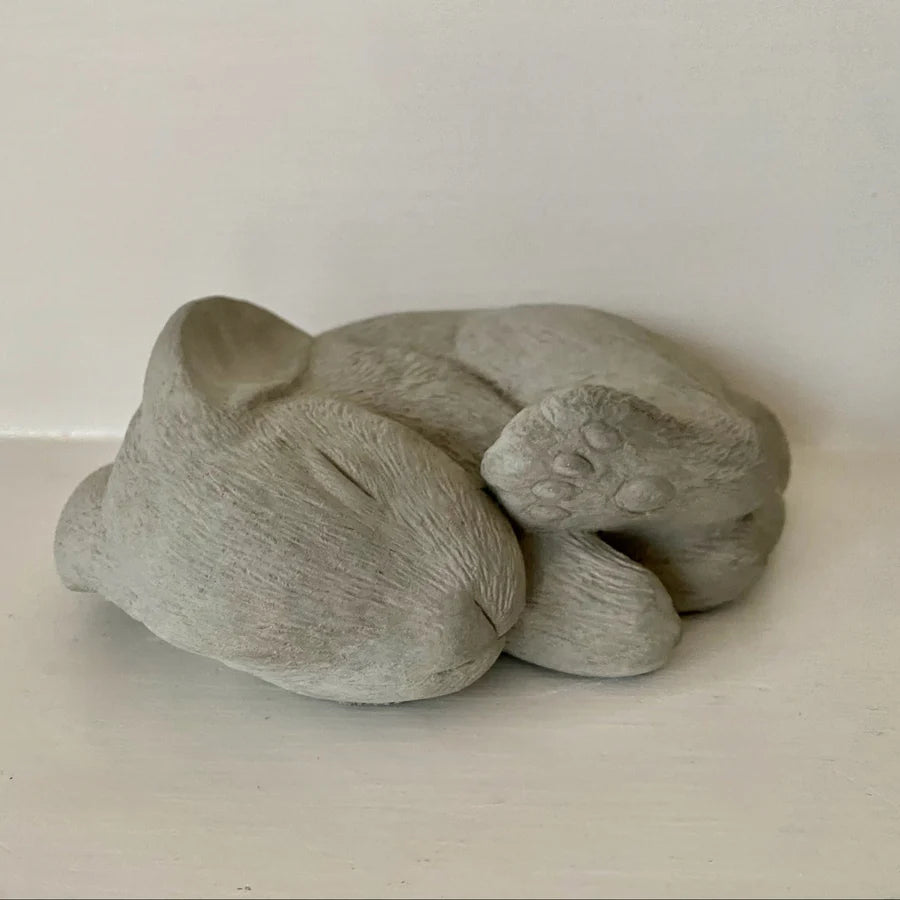 Gray concrete sculpture of a sleeping bunny curled up, handmade garden ornament, on a light surface with a neutral background