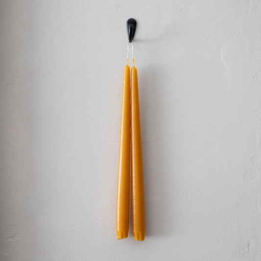 A pair of harvest gold taper candles hanging from a black hook on a white wall.