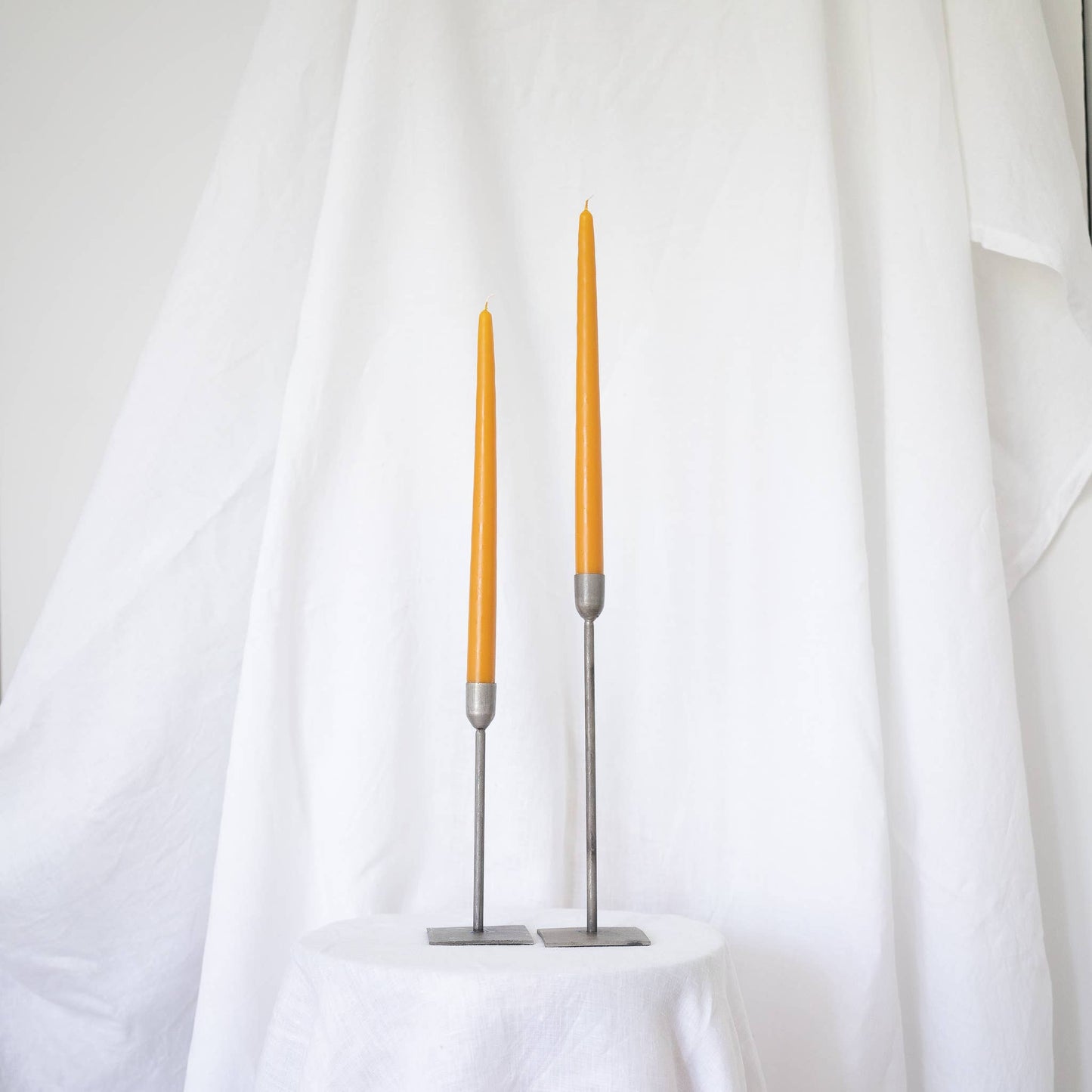 A pair of harvest gold taper candles in taper holders on a white draped wall.