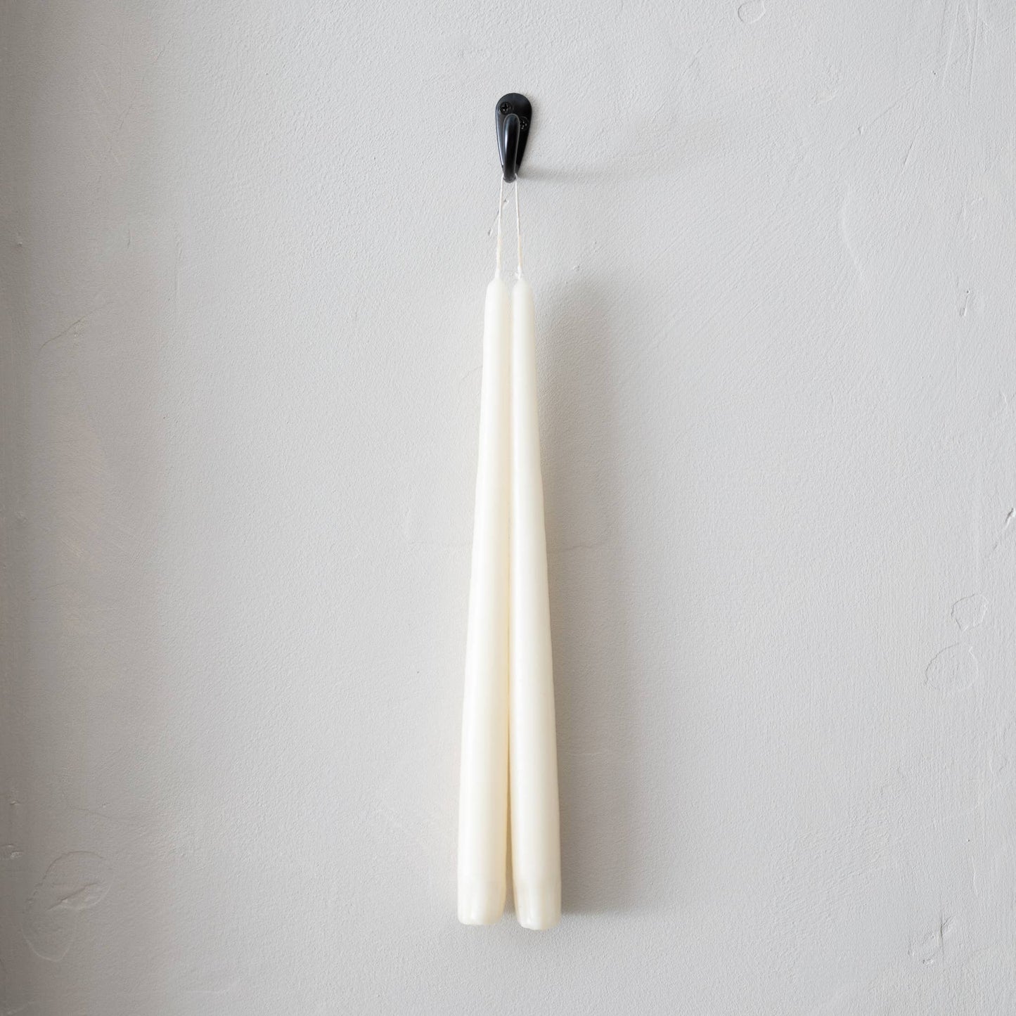 A pair of ivory taper candles hanging from a grey wall, with a black wick hanger at the top.