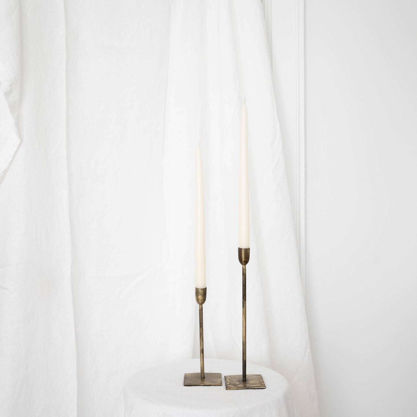 A pair of ivory taper candles in taper holders on a white draped wall.
