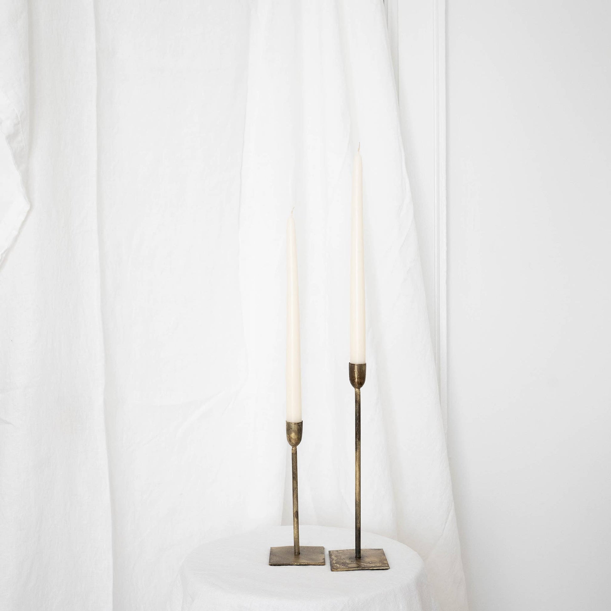 A pair of ivory taper candles in taper holders on a white draped wall.