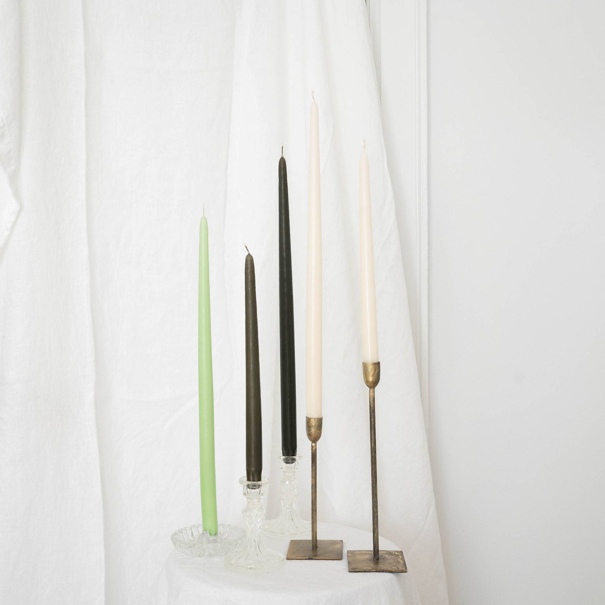 A mix of taper candles pairs in taper holders, in various colors : ivory, olive and light green, on a white draped wall.