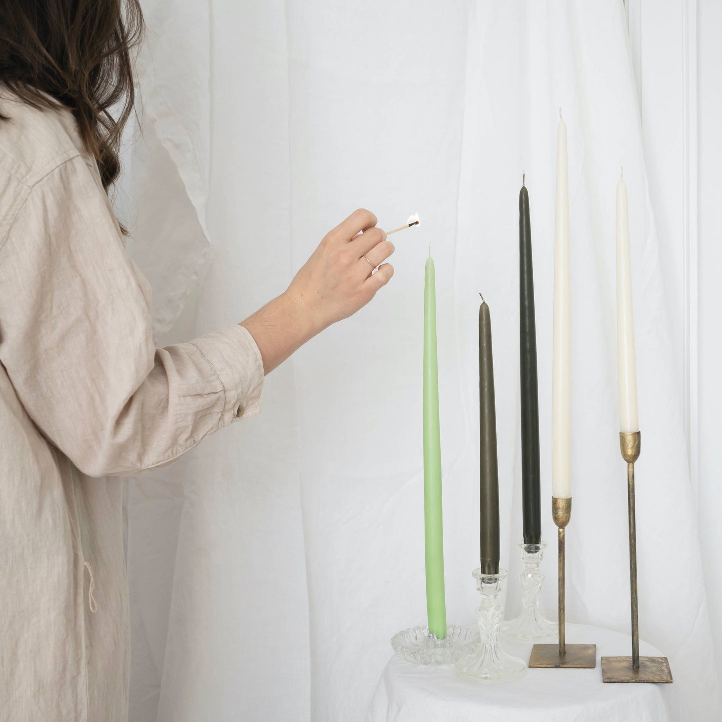 A mix of taper candles pairs in taper holders, in various colors : ivory, olive and light green, on a white draped wall. Someone is lighting one of the candle.