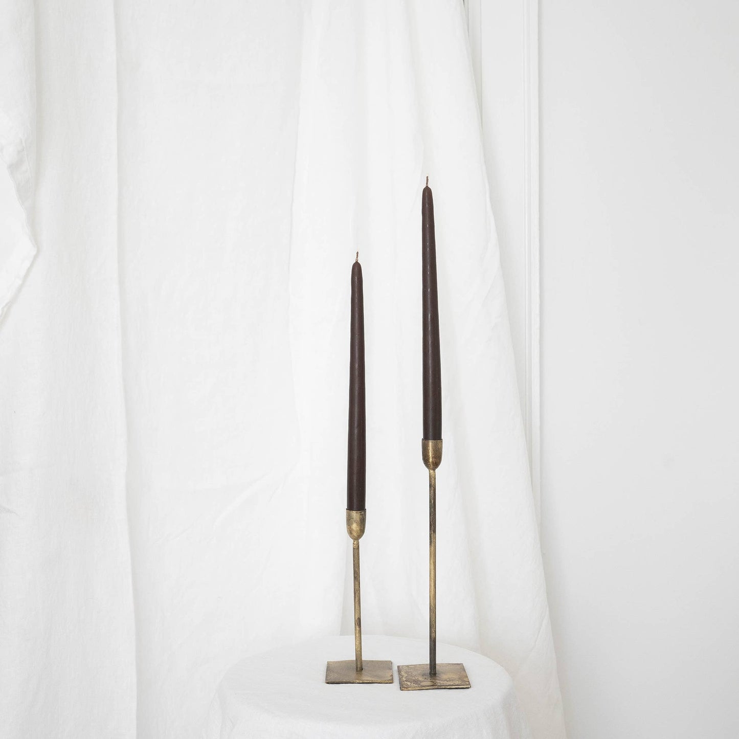 A pair of olive taper candles in taper holders on a white draped wall.