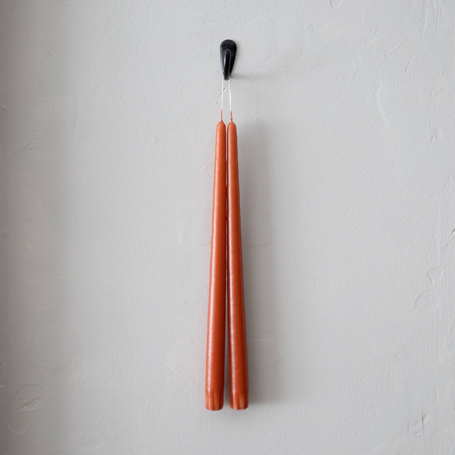 A pair of terracotta taper candles hanging from a black hook on a white wall.