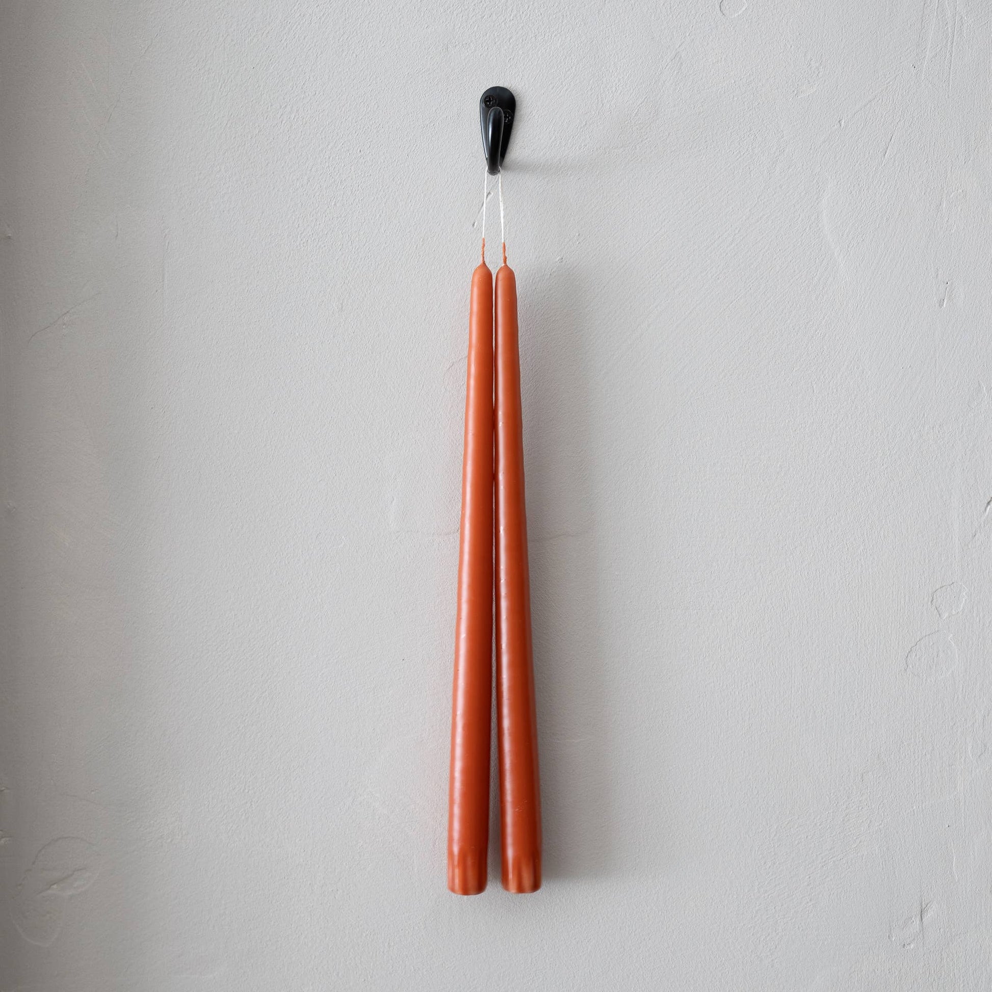 A pair of terracotta taper candles hanging from a black hook on a white wall.