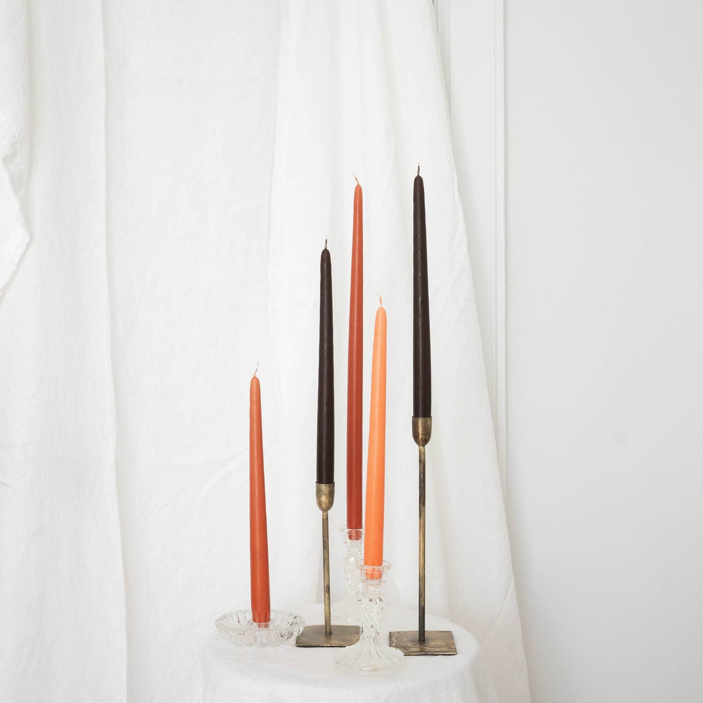 A pair of terracotta taper candles in taper holders, mixed with other taper candles, orange and brown, on a white draped wall.