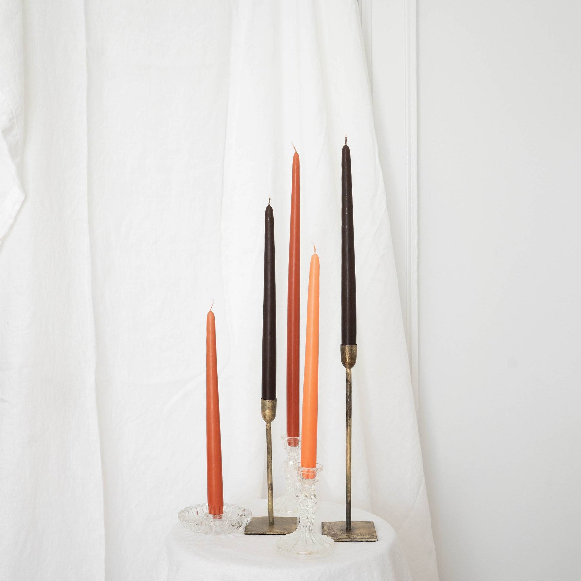 A pair of terracotta taper candles in taper holders, mixed with other taper candles, orange and brown, on a white draped wall.