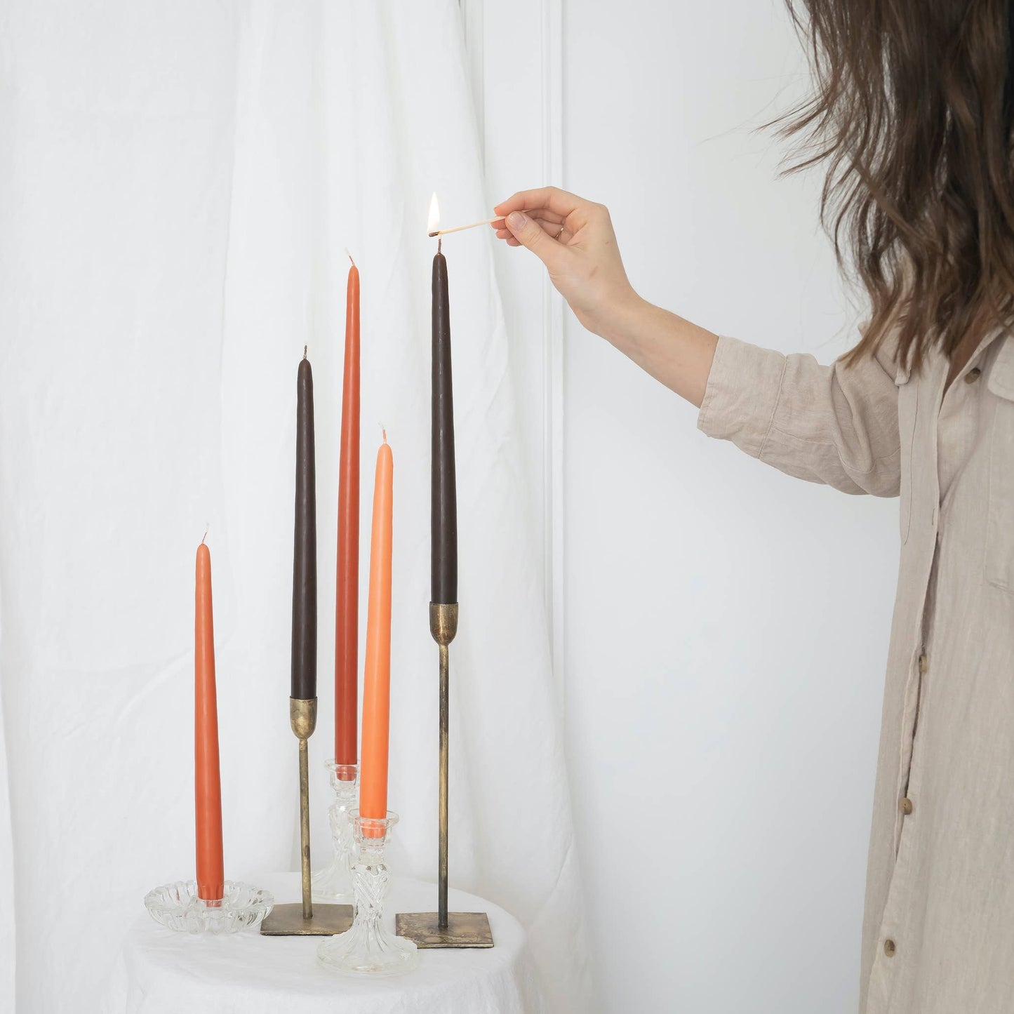 A mix of taper candles pairs in taper holders, in various colors : terracotta, orange and brown, on a white draped wall. Someone is lighting one of the candle.