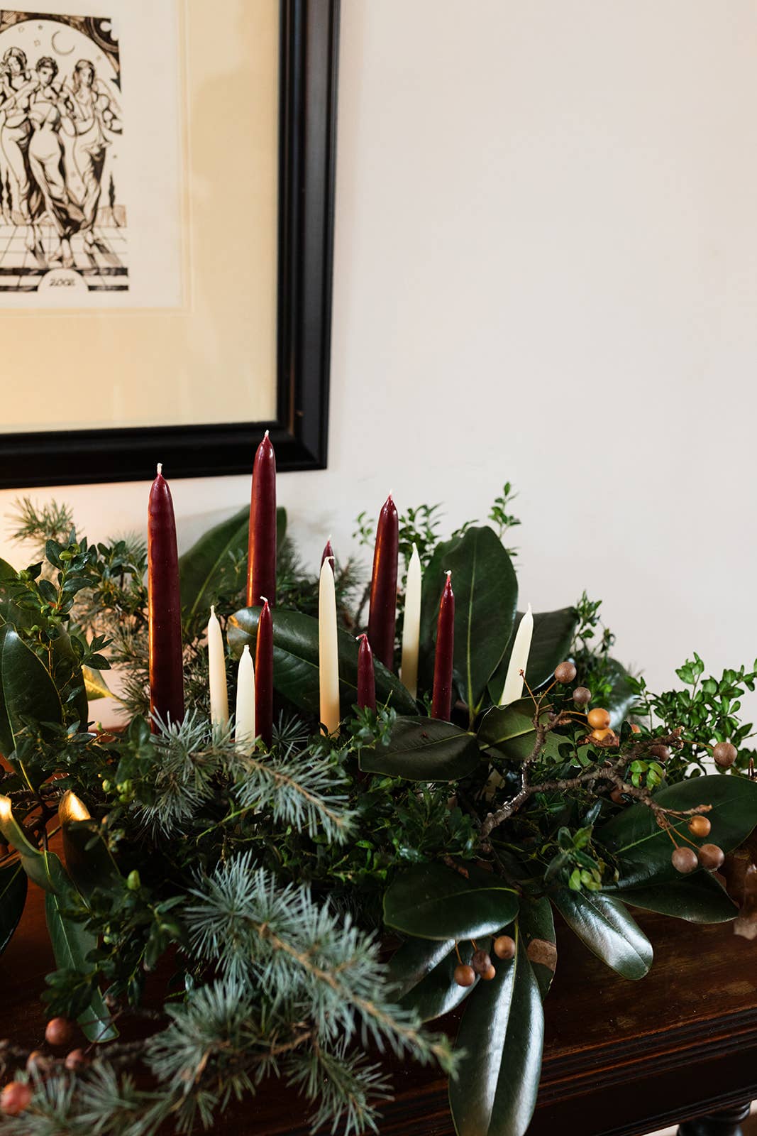 Decorative arrangement with candles and greenery on a table, framed artwork in the background.