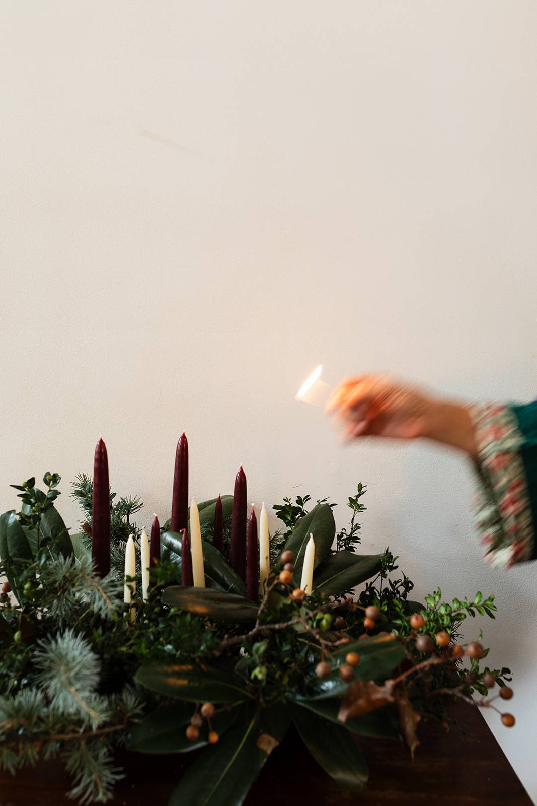 Hand lighting a candle in a decorative arrangement of candles and greenery on a white background