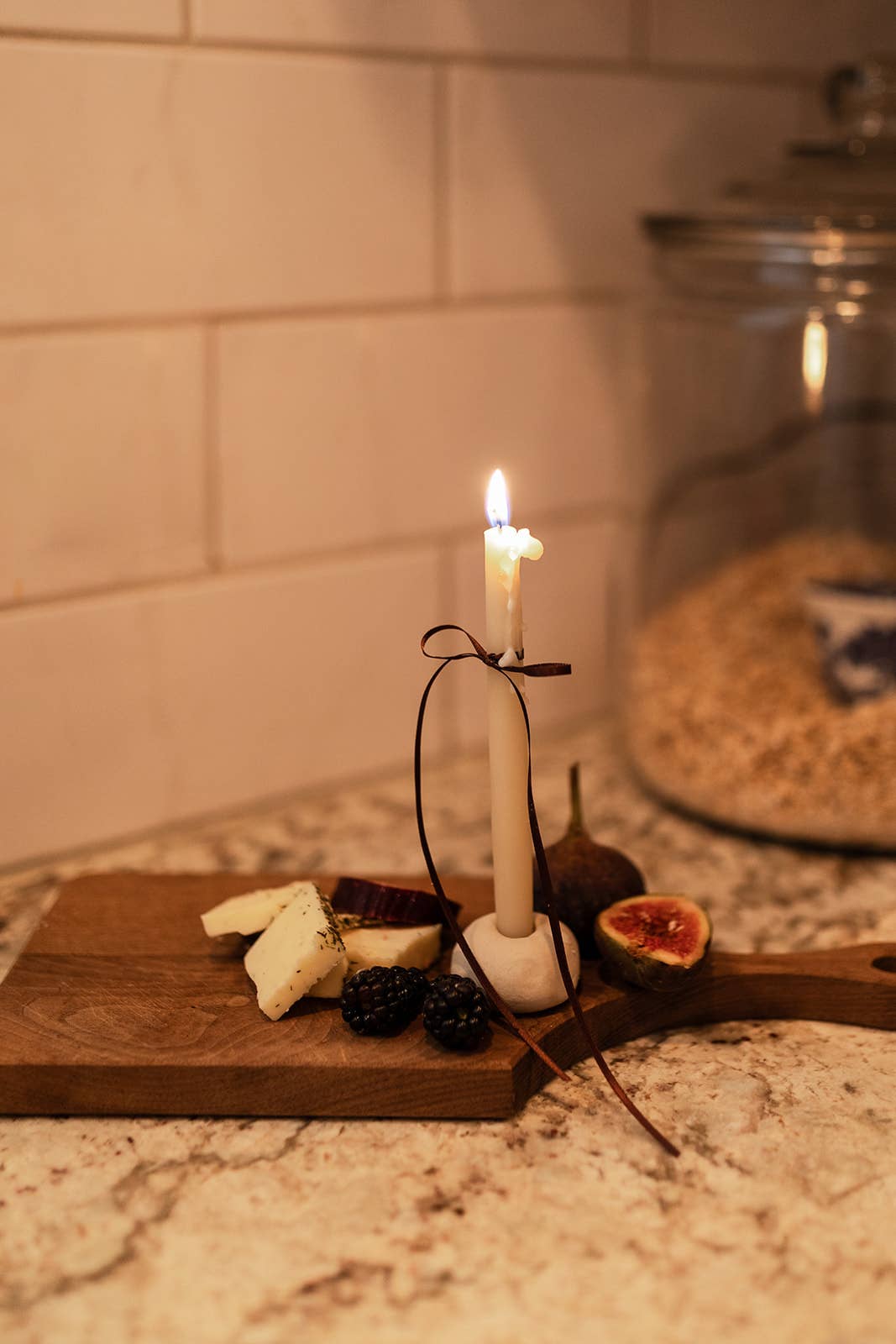 Candle on a wooden cutting board with cheese and figs against a tiled wall.