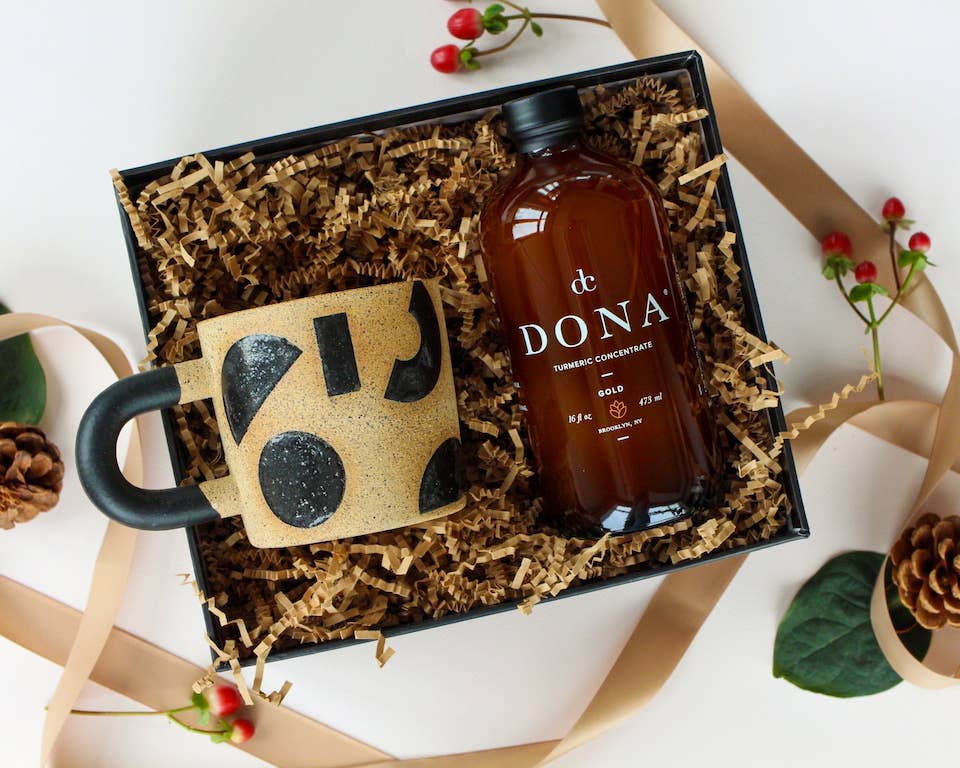 A glass bottle of Dona Turmeric Concentrate, perfect for golden lattes, with a label displaying the product name and ingredients, next to a mug with modern shapes displayed as a gift in a box. Christmas decorations are displayed in the setting.