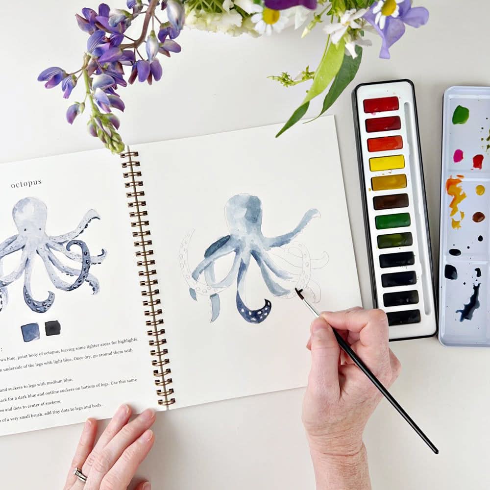Someone painting a watercolor octopus, with a watercolor paint set of 12 colors displayed in a black tin case. The lid of the tin case is used as a mixing palette. On a white background with flowers in the setting.