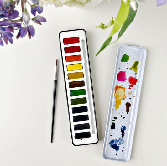 A watercolor paint set with 12 colors displayed in a black tin case alongside a round size 4 brush, with the lid of the tin case used as a mixing palette. On a white background with flowers in the setting.