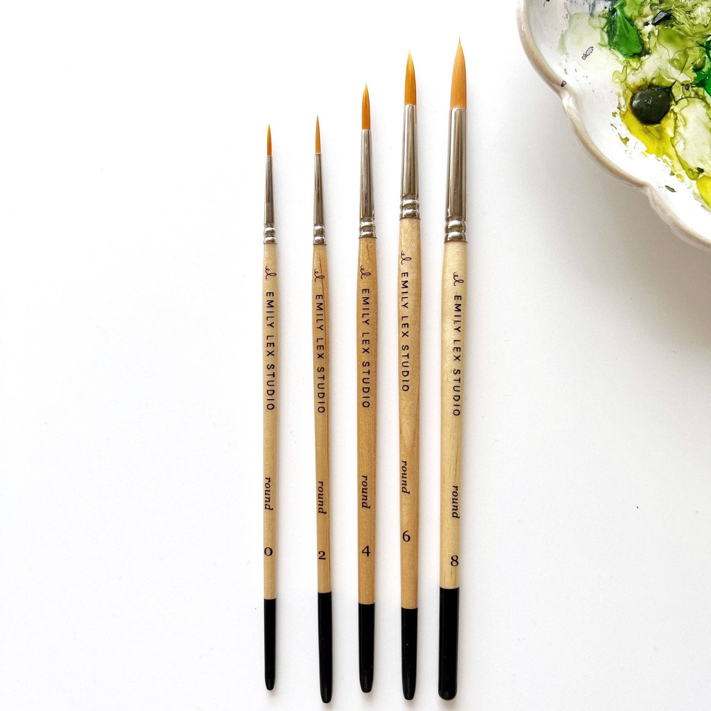 A set of five watercolor paintbrushes with golden synthetic taklon bristles, seamless aluminum ferrules, and lacquered wood handles, from emily lex studio. Next to the set is a bowl with watercolor paint.