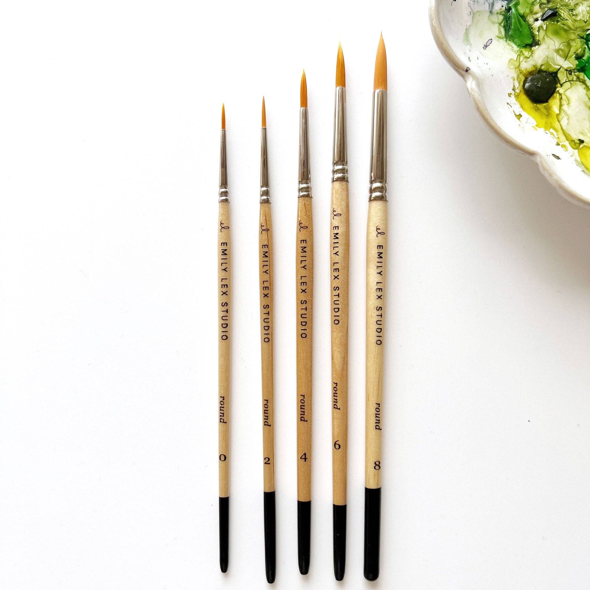 A set of five watercolor paintbrushes with golden synthetic taklon bristles, seamless aluminum ferrules, and lacquered wood handles, from emily lex studio. Next to the set is a bowl with watercolor paint.