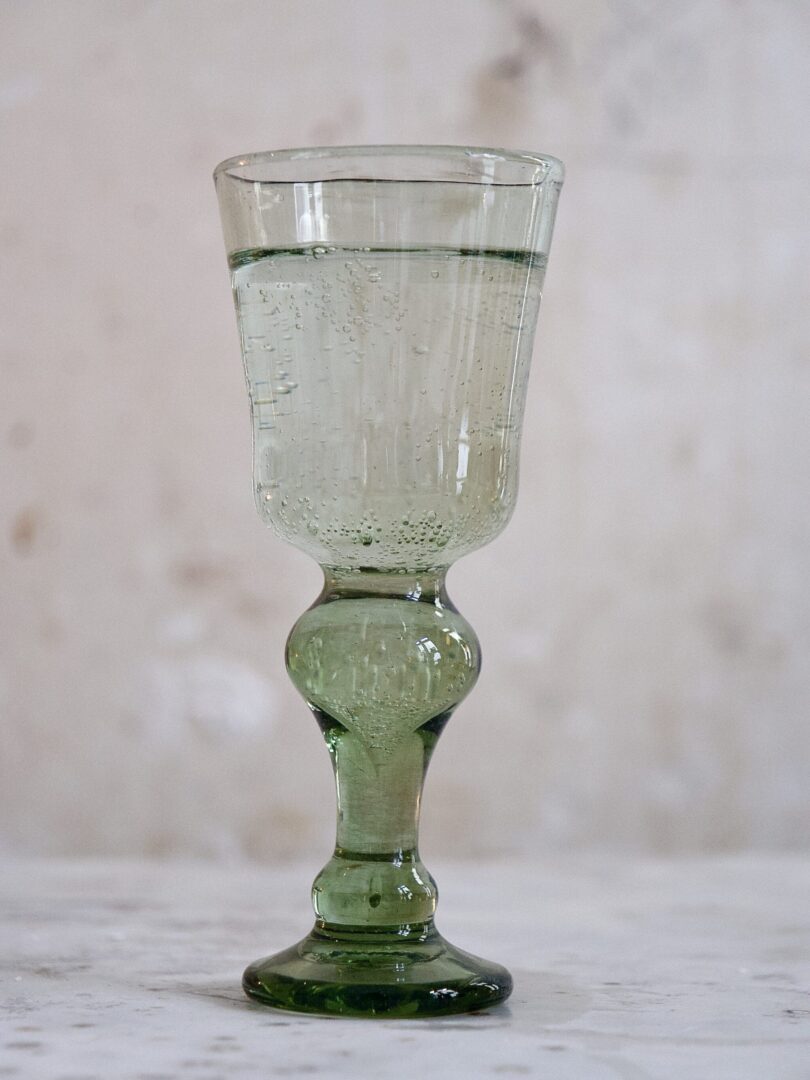 Green mouth-blown French wine glass with visible stem with transparent liquid on a marble surface with a neutral background