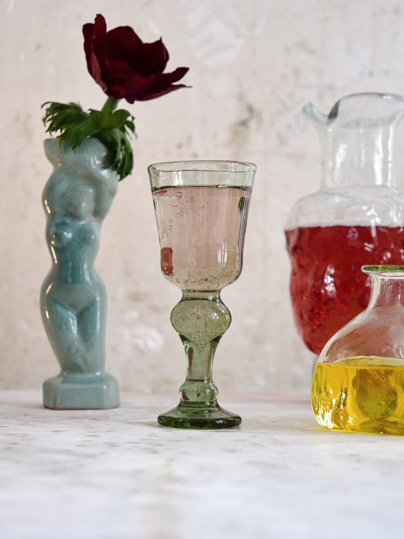 Decorative glassware including a mouth-blown green wine glass, a small vase with a rose, and a larger container with a red liquid on a marble surface.