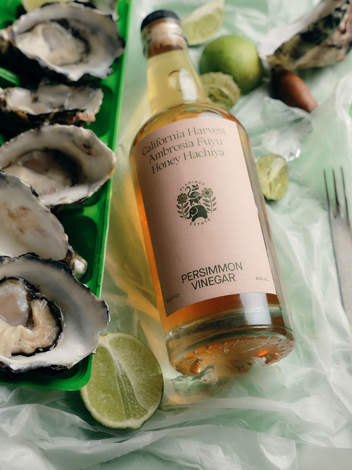 Bottle of Persimmon Vinegar, a sweet and tart Gourmet Fruit Vinegar, with oysters and lime wedges on a green surface